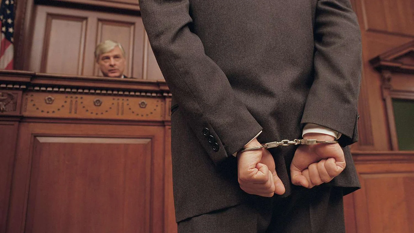 A man in a suit standing handcuffed with his back to the camera in a courtroom, a judge seated at the bench behind him