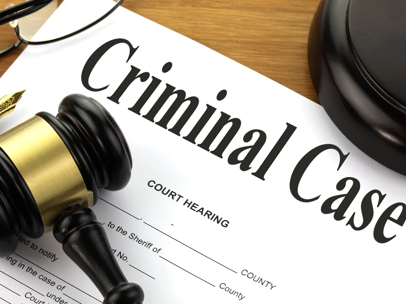 A printed "Criminal Case / Court Hearing" form on a wooden desk beside a gavel, a fountain pen and a pair of glasses.