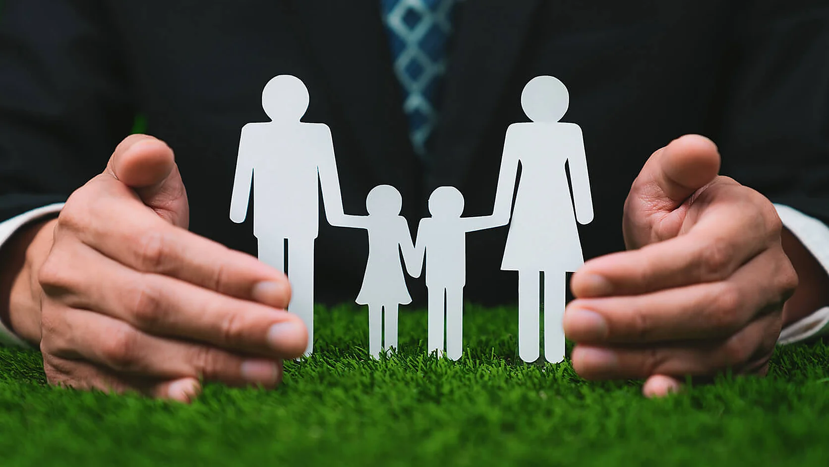 A man in a business suit cupping his hands protectively around a paper cut-out family of two adults and two children holding hands, standing on grass.