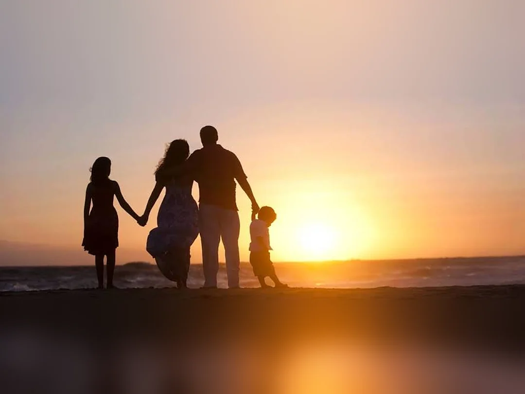 A family of four in silhouette on a beach at sunset, two parents holding hands with a daughter and a son, facing the sun over the water.