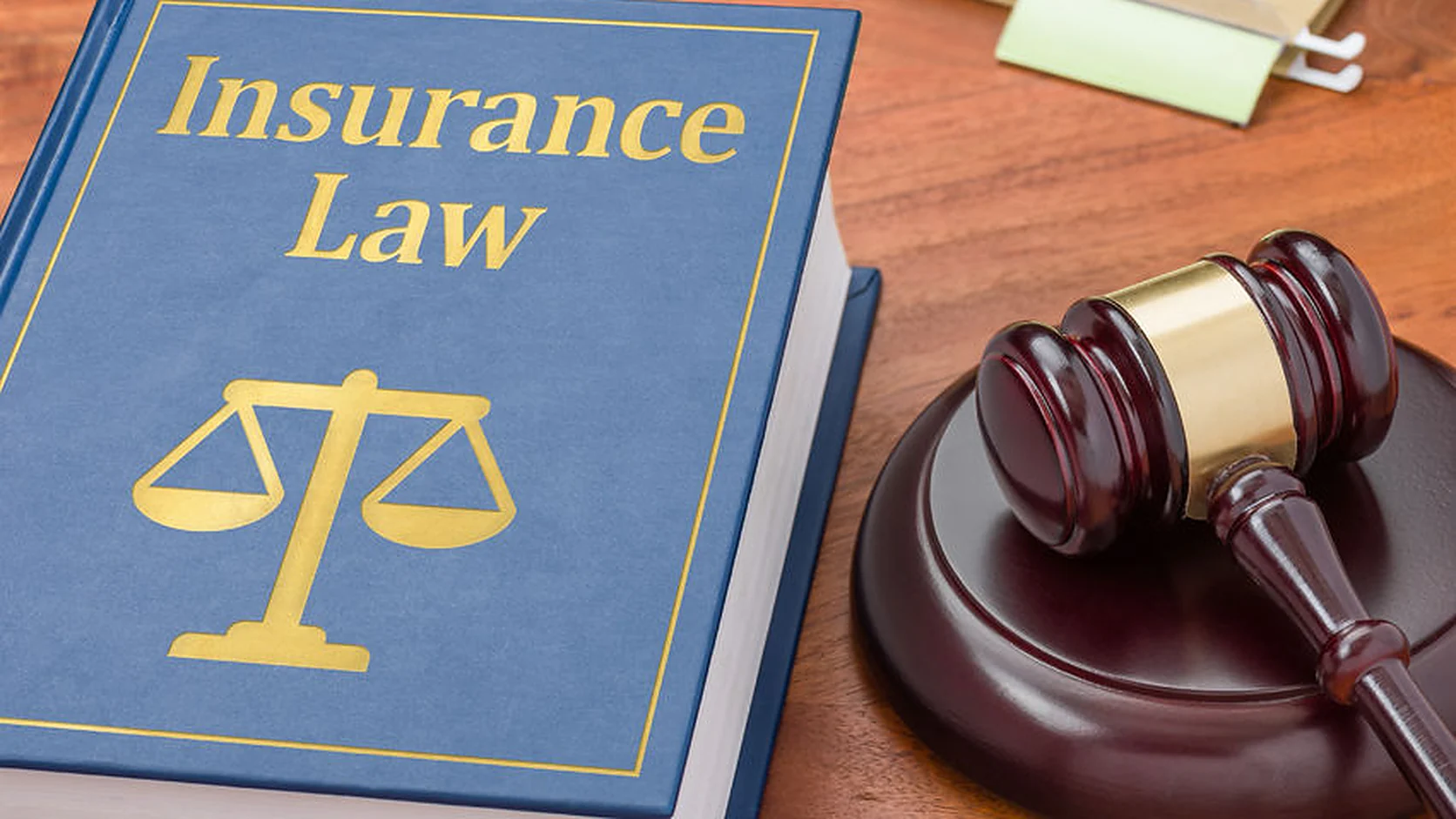 A hardback "Insurance Law" law book with a scales-of-justice emblem on its cover, lying beside a gavel and sound block on a wooden desk.